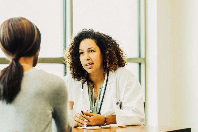 A doctor at her desk faces the camera and shares information with a female patient facing away from the camera